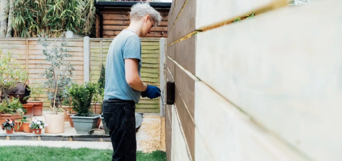 DIY fencing - young man worker paints with a roller a wooden board fence in the garden